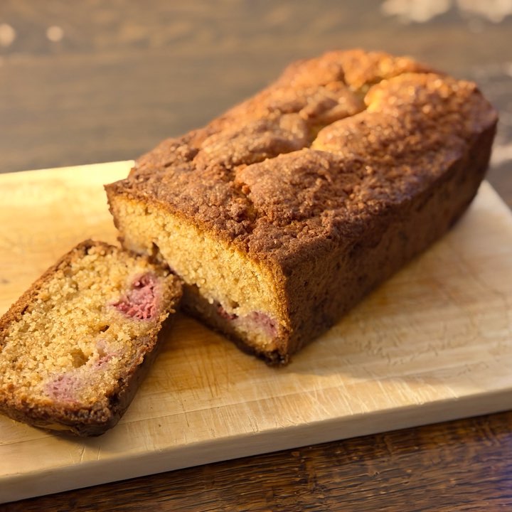 A raspberry and white chocolate loaf with a slice cut, on a wooden board.